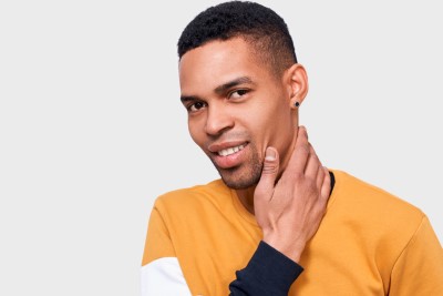 Image of young handsome African American man touching his face and neck with hand and looking to the camera while standing against white studio background. Good-looking male feeling clean and fresh.