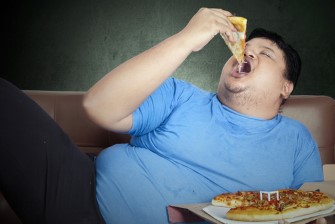 Obese,Person,Eats,Pizza,While,Sitting,On,Couch,At,Home